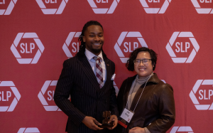 Two people with an award in front of a red backdrop.